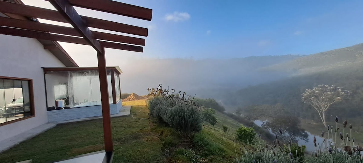 A serene outdoor view showcases the house's modern design, with large glass windows reflecting the natural landscape. Lush greenery extends across the foreground, while mist gently envelops the distant hills, providing a tranquil ambiance.