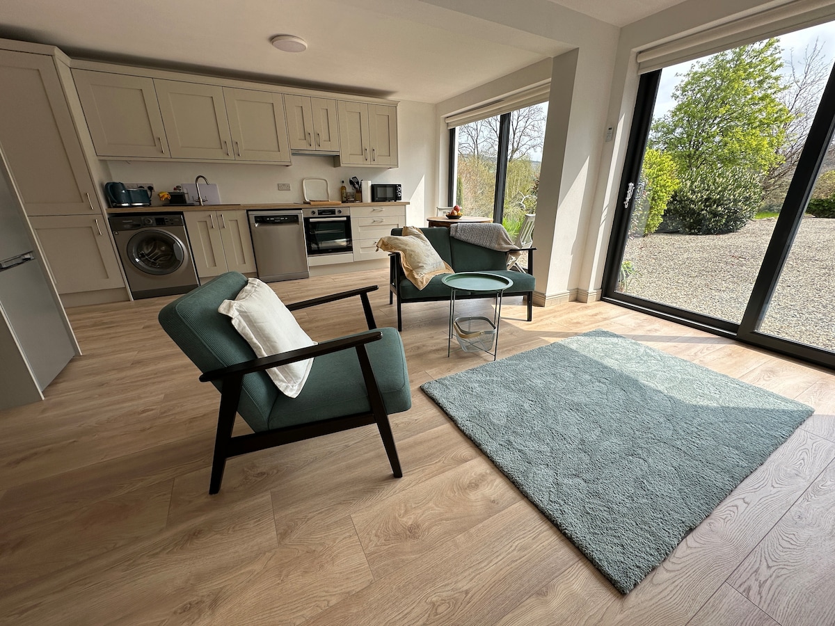 A bright kitchen area features light cabinetry and modern appliances. Two comfortable chairs are positioned near a small glass table, with a soft area rug enhancing the space. Large windows allow natural light to fill the room, providing views of the surrounding grounds.