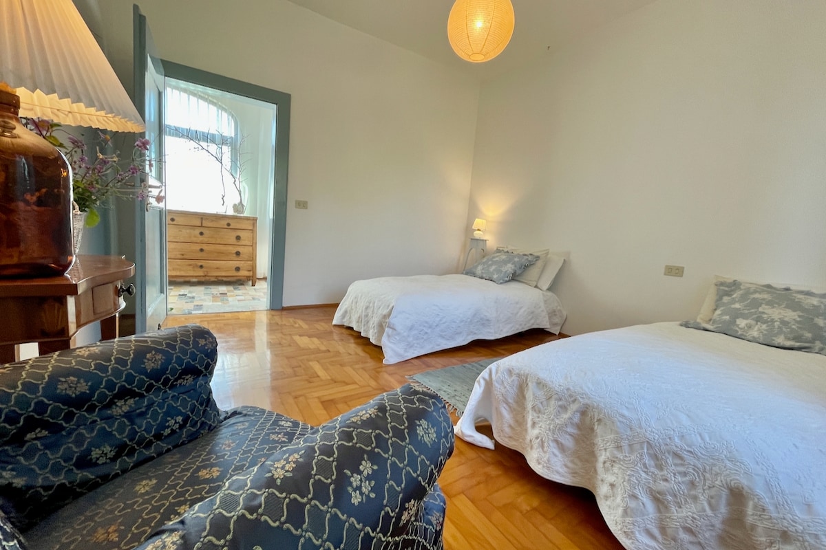 The guest room features two spacious beds with white bedding, complemented by soft pillows. A cozy armchair is positioned nearby, and a wooden dresser is visible in the background. Natural light illuminates the space, enhancing its inviting atmosphere.