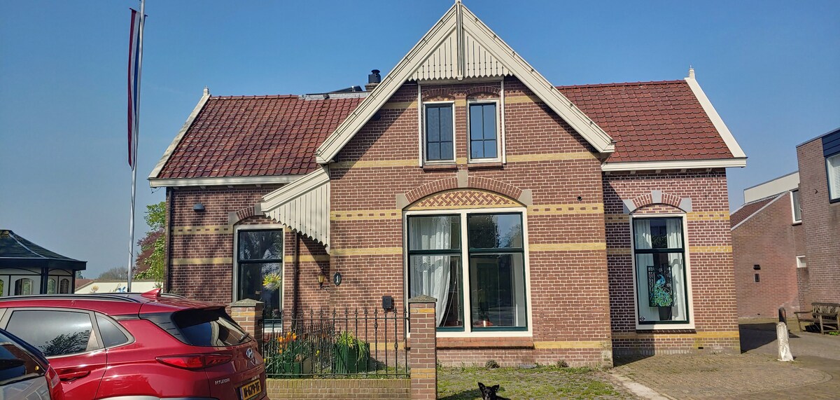 The exterior of a well-maintained brick house is showcased, featuring distinct architectural details such as arched windows and a decorative awning. A manicured yard with flowers is visible, while a red car is parked nearby. Clear blue skies create a serene backdrop.
