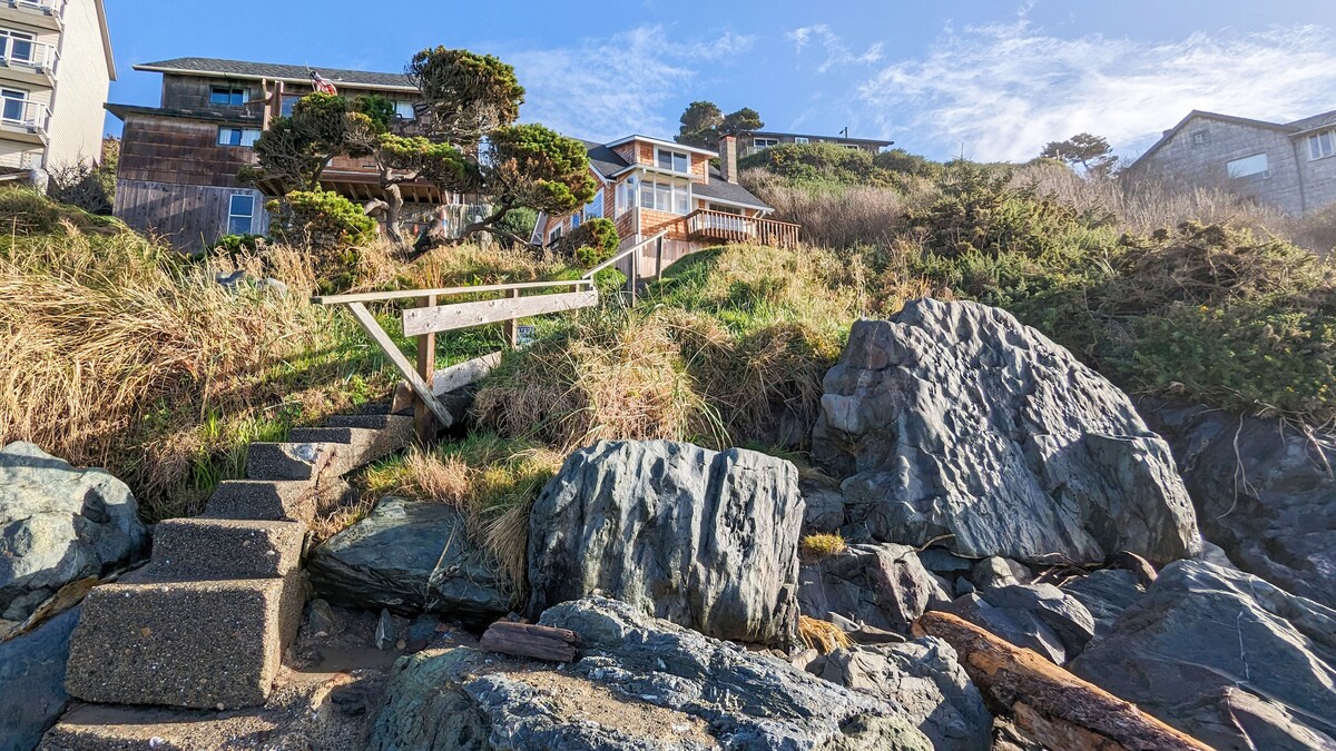 A pathway made of stone steps leads from rocky terrain up towards a cabin set on a hillside, surrounded by lush grass and coastal vegetation. The structure features large windows and a wooden deck, highlighting its beachside location under a clear blue sky.