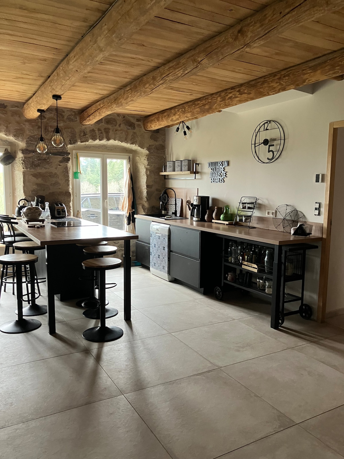 The modern kitchen features wooden beams and stone walls, enhancing its rustic charm. A large island with bar stools provides ample workspace. A variety of kitchen appliances are neatly arranged, and natural light floods in through a window, illuminating the sleek countertops.