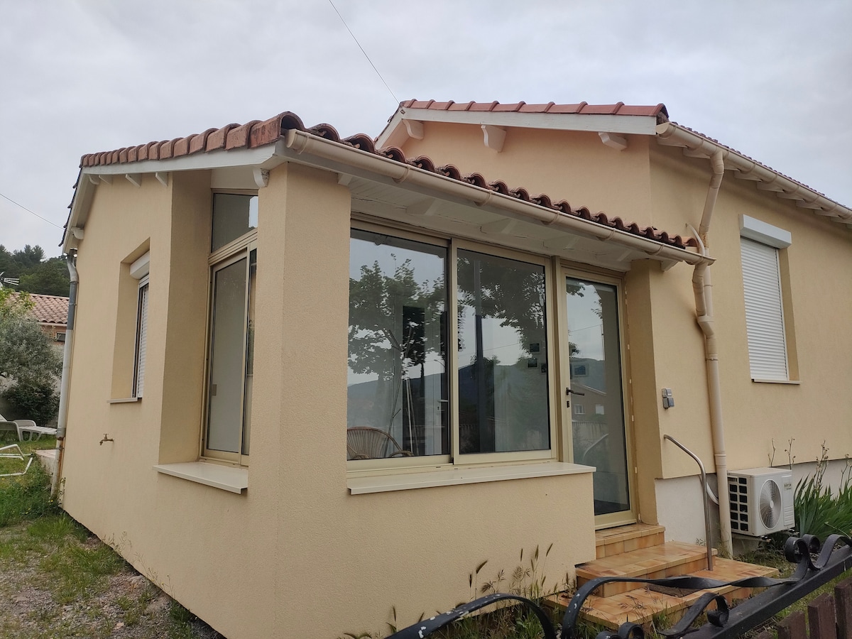 A single-story house features large windows that invite natural light into a bright, open space. The exterior is finished with light-colored stucco, and a tiled roof adds character. A small outdoor area is visible, suggesting opportunities for relaxation and enjoyment.