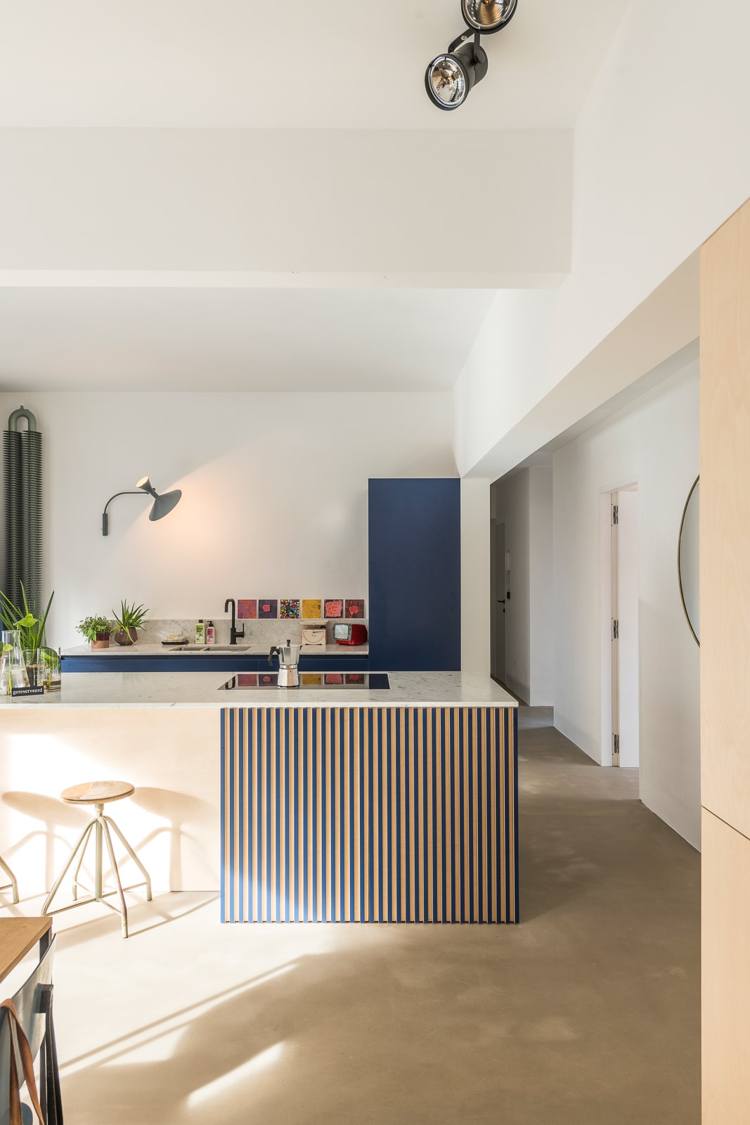 An open kitchen area is highlighted by a striped kitchen island, featuring a modern cooktop. Natural light filters through a nearby window, while plants add a touch of greenery. The space is designed to be functional and visually appealing.