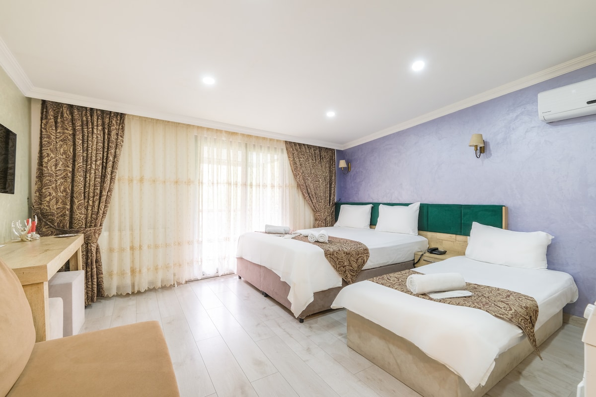 A spacious room features two beds with light linens and decorative throws. A large window with sheer curtains allows natural light to fill the space. A wooden desk and chair are positioned near the window, while a wall-mounted television is visible. Soft lighting enhances the calming atmosphere.