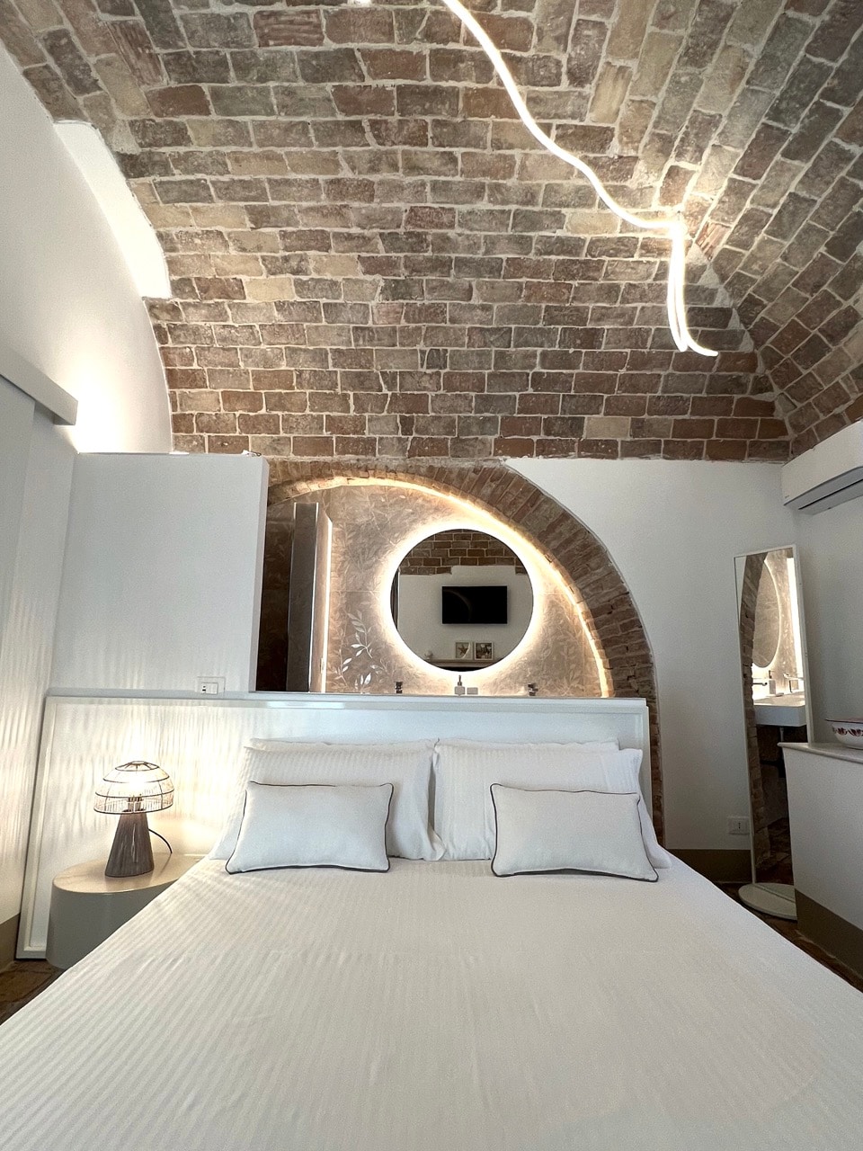 A spacious sleeping area features a comfortable bedding setup with two decorative cushions and a sleek lamp beside the bed. An arched stone wall complements the unique ceiling design, while a circular mirror adds depth to the ambiance. Natural light filters softly through the window.