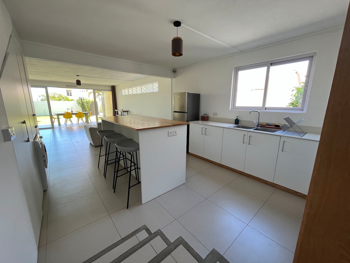 A modern kitchen area features minimalistic white cabinetry and a wooden countertop, complemented by bar stools for seating. Natural light floods in from a large window, illuminating the open layout that connects to a dining space with yellow chairs, creating a bright and spacious environment.