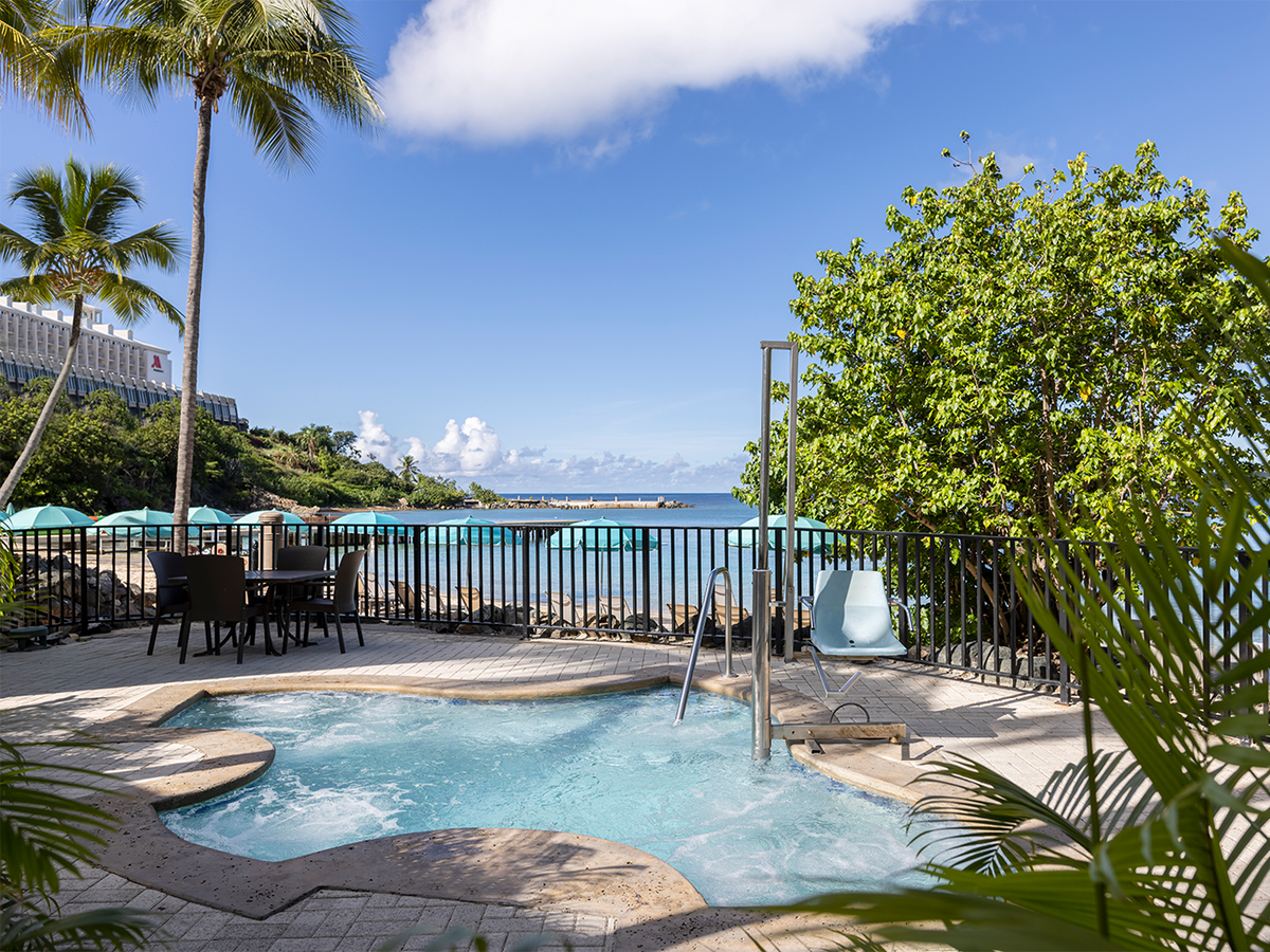 A whirlpool spa is surrounded by tropical landscaping, with palm trees and lush greenery framing the view. The sparkling water is visible, reflecting the blue sky. A seating area with dark wicker chairs and a table offers a space for relaxation near the water's edge.