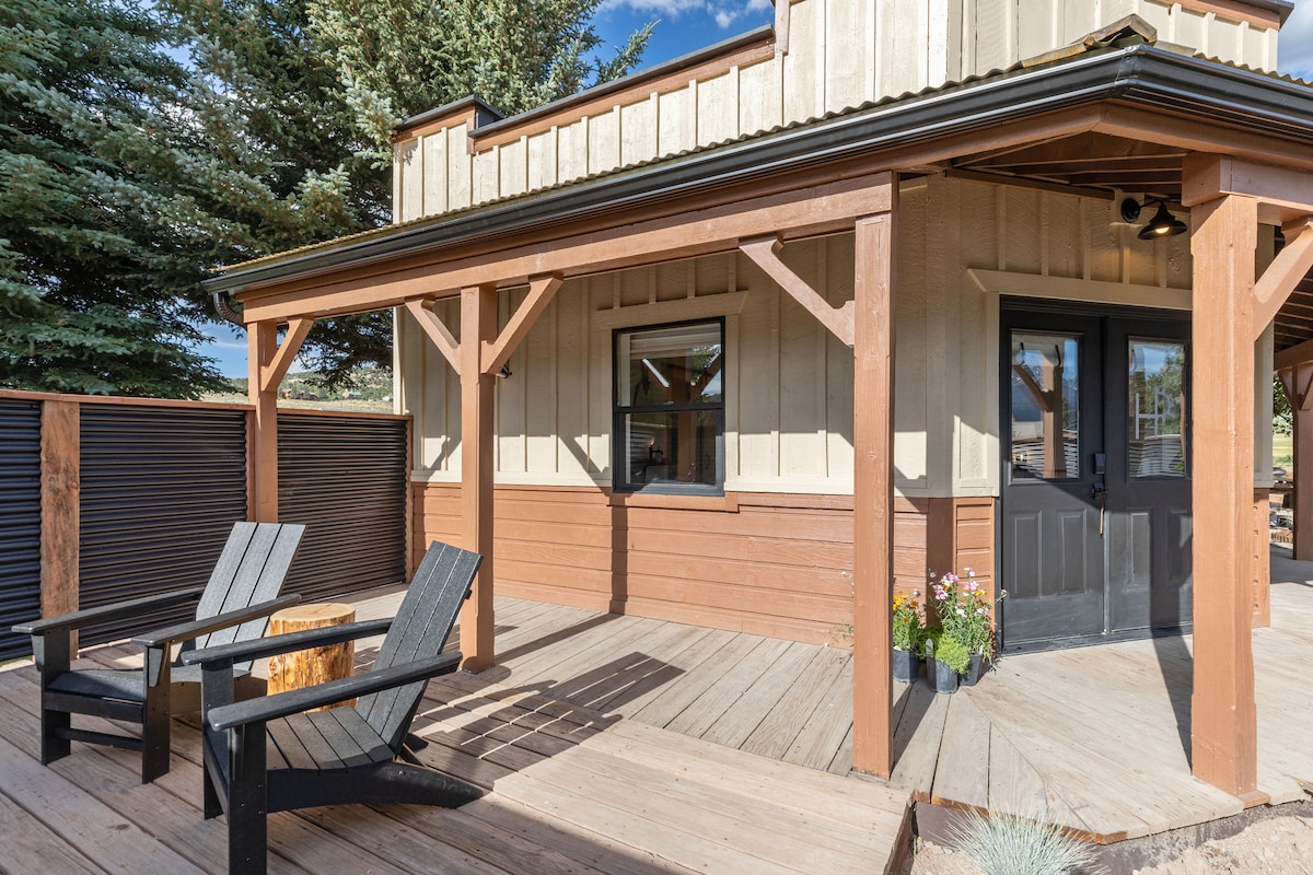 A welcoming entry features a covered patio with two black Adirondack chairs positioned next to a wooden fire pit. The surrounding deck is made of weathered wood, and the charming exterior includes neutral siding with brown accents and a front door. Potted flowers add a touch of color.