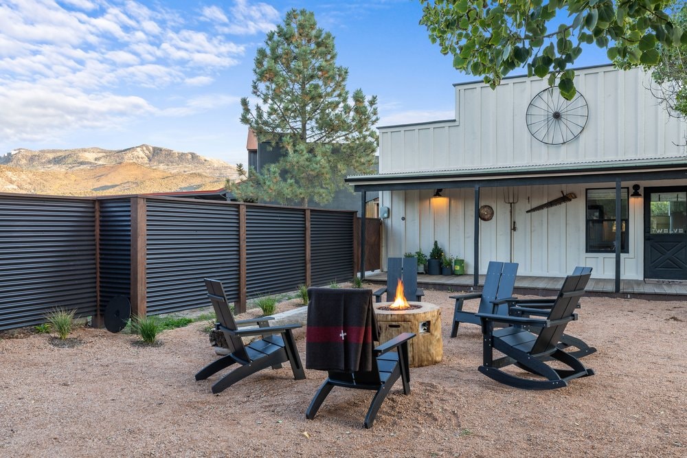 An outdoor seating area is arranged around a fire pit, featuring several black adirondack chairs positioned on a gravel surface. A rustic wooden stump serves as a table, and a blanket is draped over one chair. Natural surroundings include trees and a view of distant mountains.