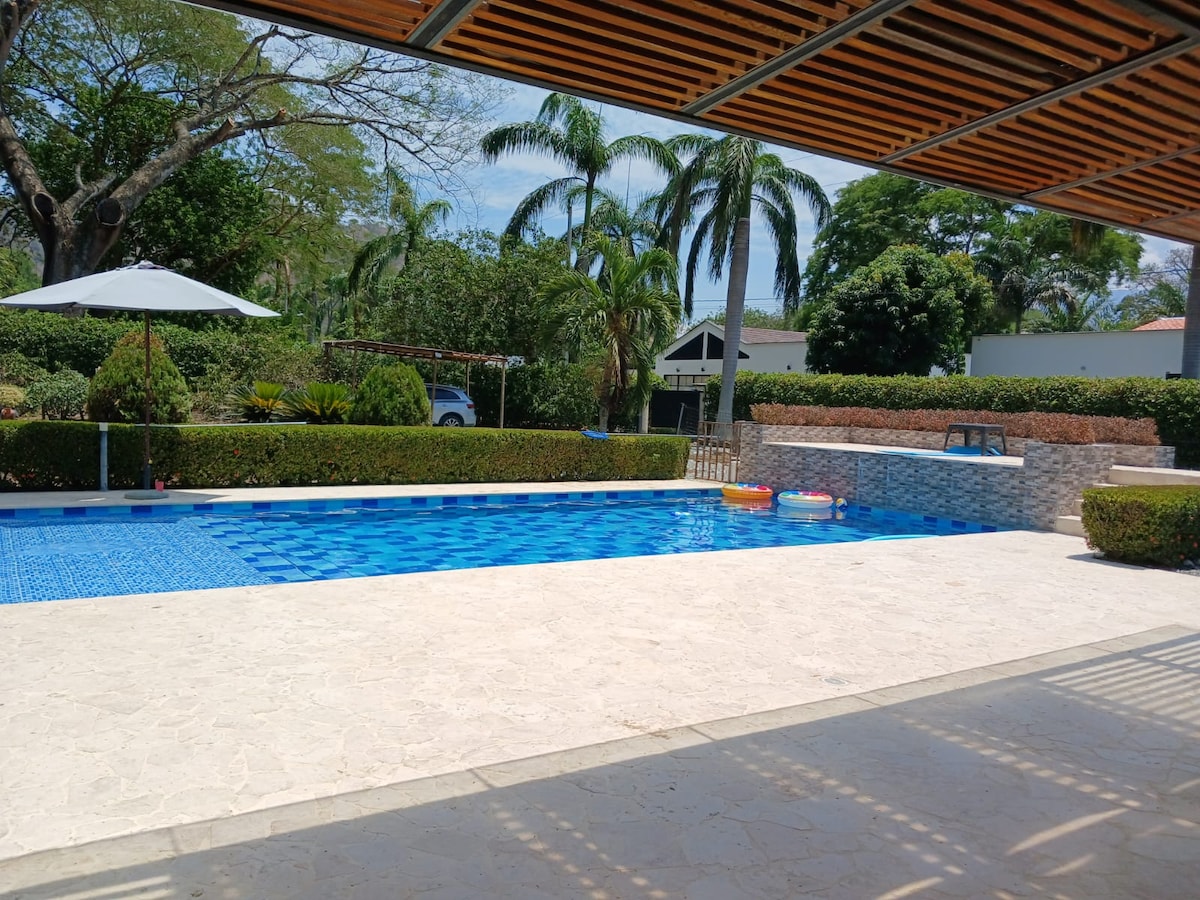 An outdoor swimming pool is visible, featuring clear blue water and surrounded by lush greenery. Sun loungers and umbrellas are arranged nearby, with a shaded area for relaxation. The pool is bordered by elegantly tiled surfaces, and colorful inflatable floats are resting on the water.