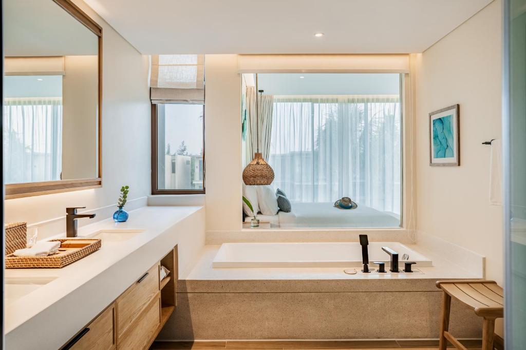 A modern bathroom is featured, showcasing a freestanding bathtub near a large window. Natural light illuminates the space, with a minimalist vanity composed of light wood and white surfaces. Decorative items are placed on the countertop, and a small green plant adds a touch of nature.