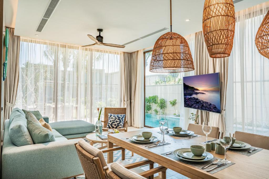 A light-filled living and dining area features a spacious sofa adorned with accent cushions. A wooden dining table is set for four, complemented by natural woven pendant lights. Large windows frame a view of the garden pool, enhancing the open-air concept.