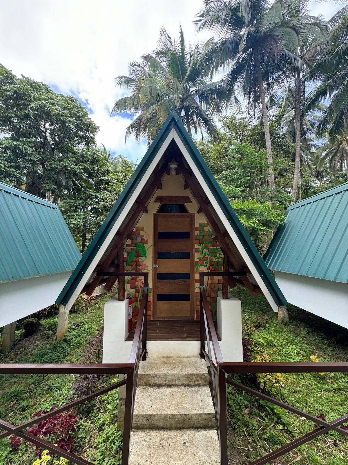 An A-frame structure with a colorful mural at the entrance is surrounded by lush greenery and palm trees. Stone steps lead up to the door, which features multiple panels and a warm wood finish. The sloping roof is complemented by blue accents.