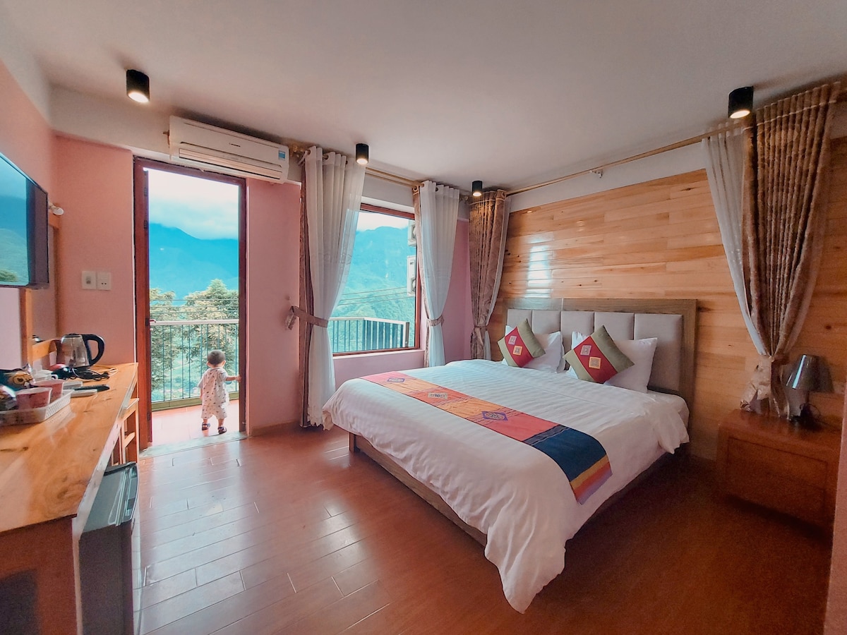 A spacious bedroom features a large bed with decorative pillows and a wooden accent wall. Natural light fills the room through sliding glass doors that lead to a balcony with mountain views. A child is seen looking out, while a small desk and appliances are present.