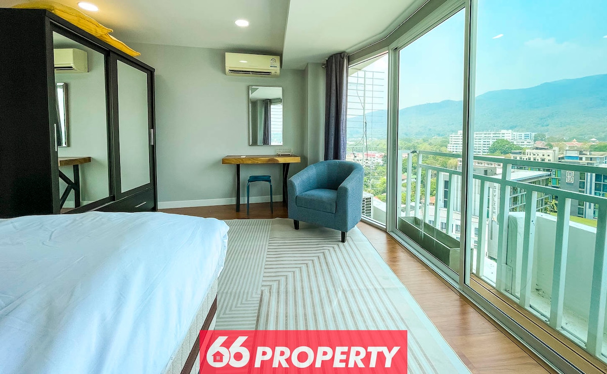 A bright and airy room features a spacious bed, a compact desk with a mirror, and a cozy blue chair. Floor-to-ceiling windows provide expansive views of the mountains and cityscape, while soft light fills the space. A neutral rug adds texture to the wooden floor.