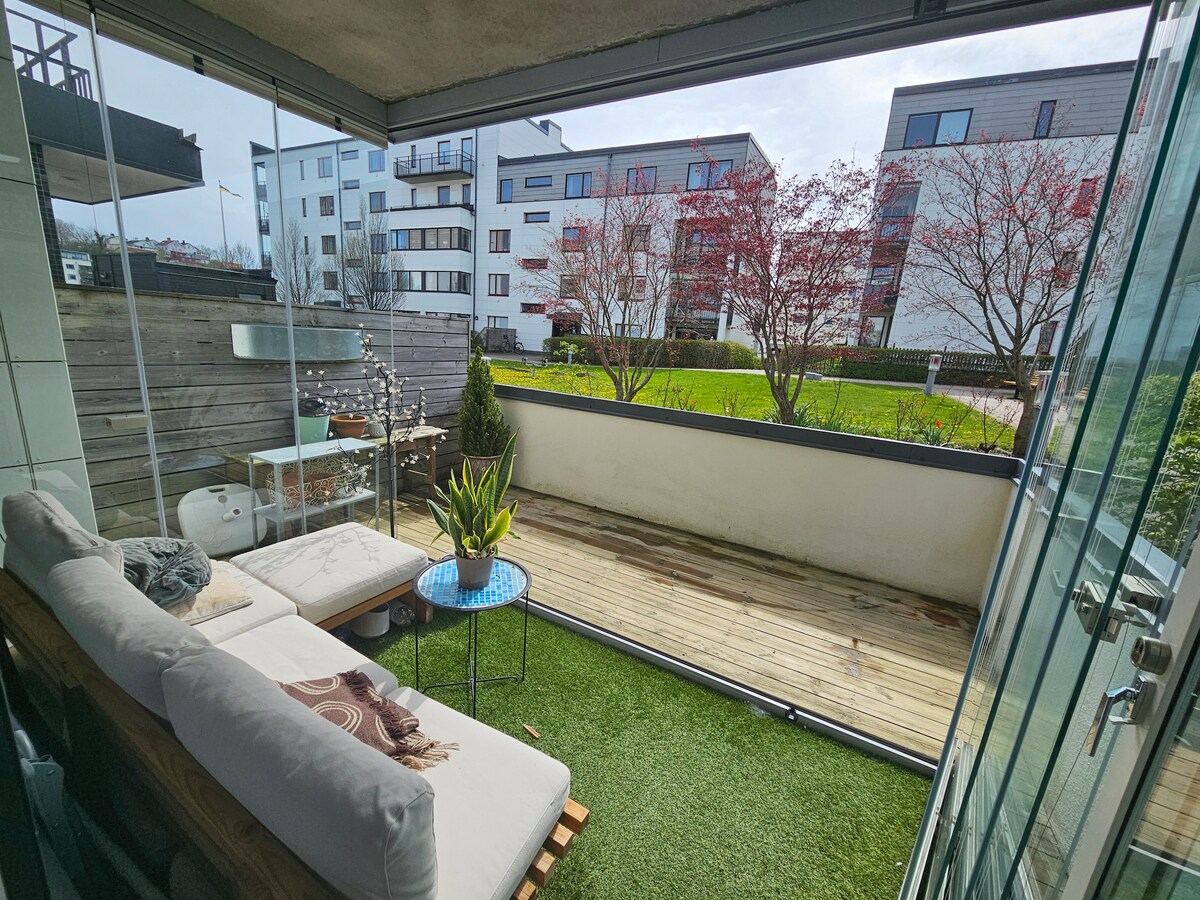 An outdoor balcony area features a cozy seating arrangement with a soft cushion on a wooden frame. A small blue table is positioned in front of a green potted plant, with soft grass underfoot. Greenery and buildings are visible in the background.