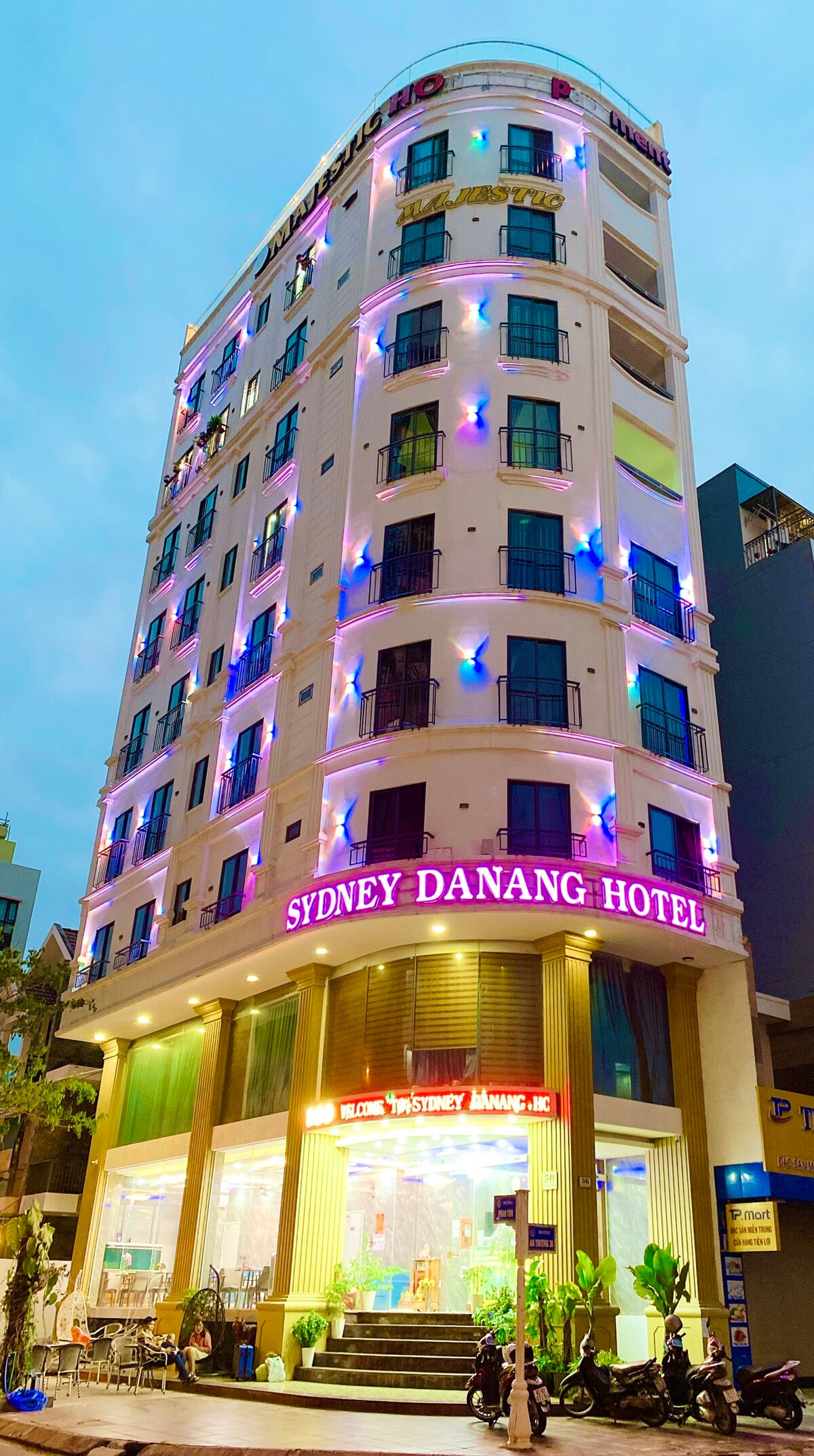 The hotel exterior features a modern design with multiple floors, illuminated by colorful lights. Large windows offer glimpses of the interior. The entrance is framed by green plants, providing a welcoming touch to guests arriving at Sydney Da Nang Hotel.