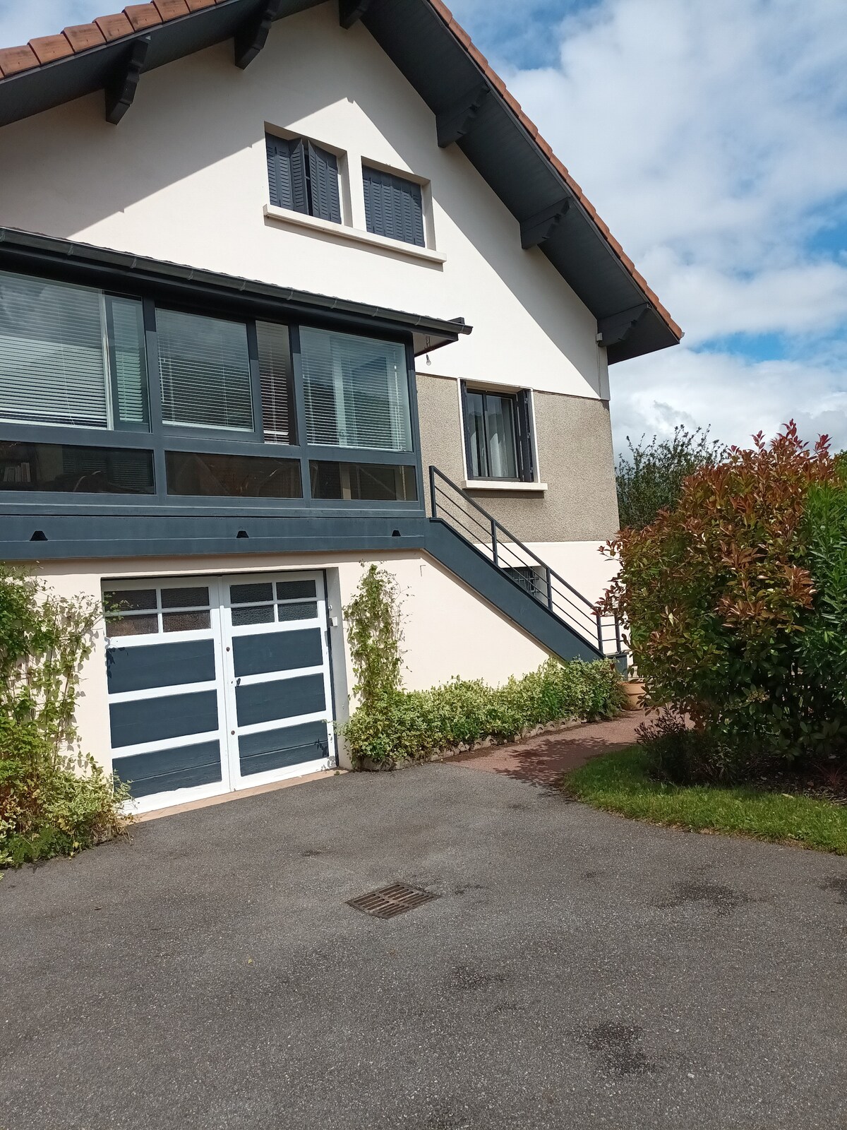 A two-story villa is depicted, featuring a combination of modern and traditional architecture. The entrance includes a large glass wall with multiple windows. A set of stairs leads to the second floor, complemented by greenery along the driveway and a garage door visible at ground level.
