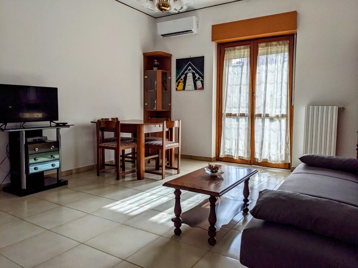 A bright living area features a wooden table with four chairs, complemented by a cozy gray couch. A television is positioned on a black stand. Natural light enters through two windows adorned with light curtains, enhancing the spacious feel of the room.