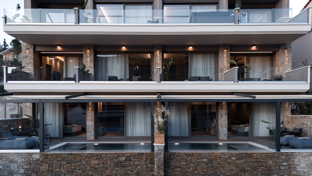 The exterior of a multi-level building showcases a blend of modern architecture and natural stone. Large glass doors open to elegantly decorated spaces, with potted plants visible. A serene pool reflects the surrounding area, enhancing the inviting atmosphere of the property.