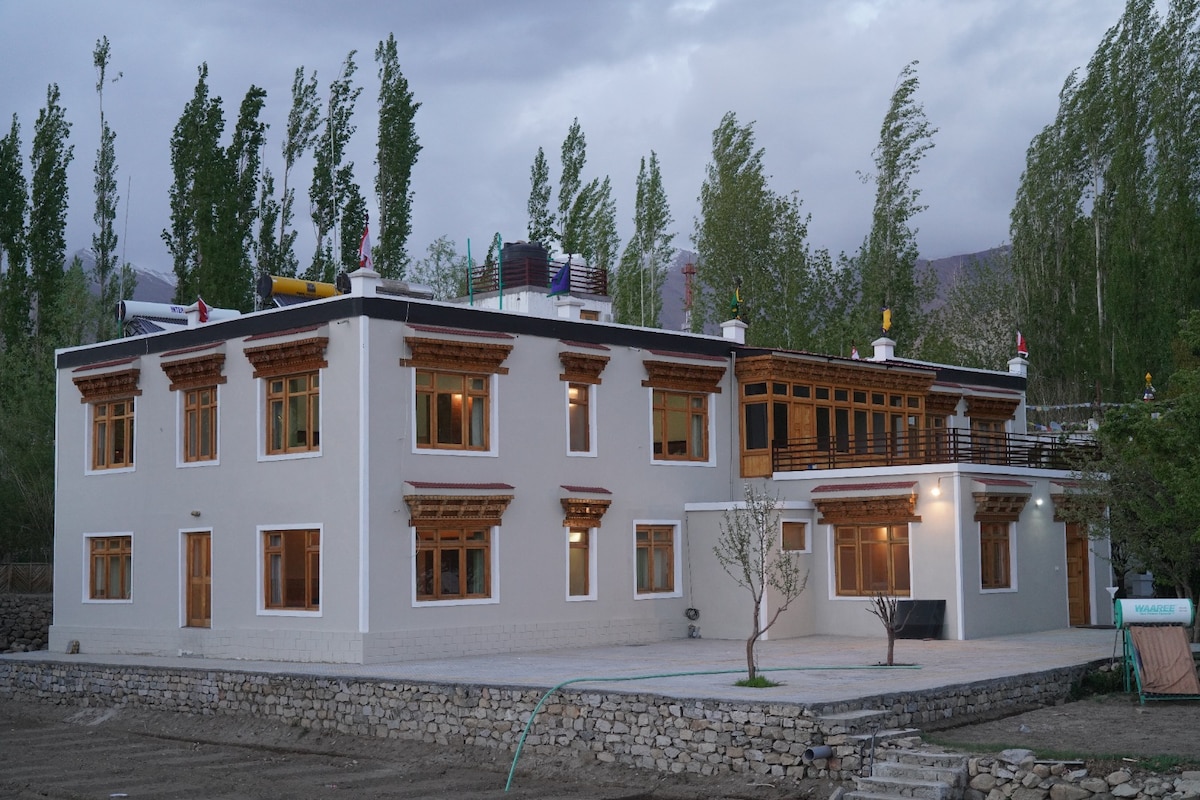 The homestay features a two-story building constructed in traditional Ladakhi style, characterized by wooden floors and ornate window frames. Surrounding greenery and poplar trees enhance the natural setting, while a gravel path leads to the entrance, creating an inviting atmosphere.