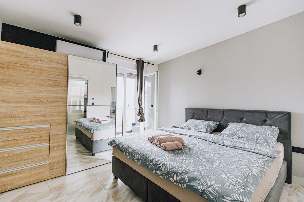 A private room features a spacious bed with floral-patterned linens, complemented by a large mirror that reflects the room's decor. Natural light enters through glass doors that lead to a terrace, while a wooden wardrobe provides ample storage.