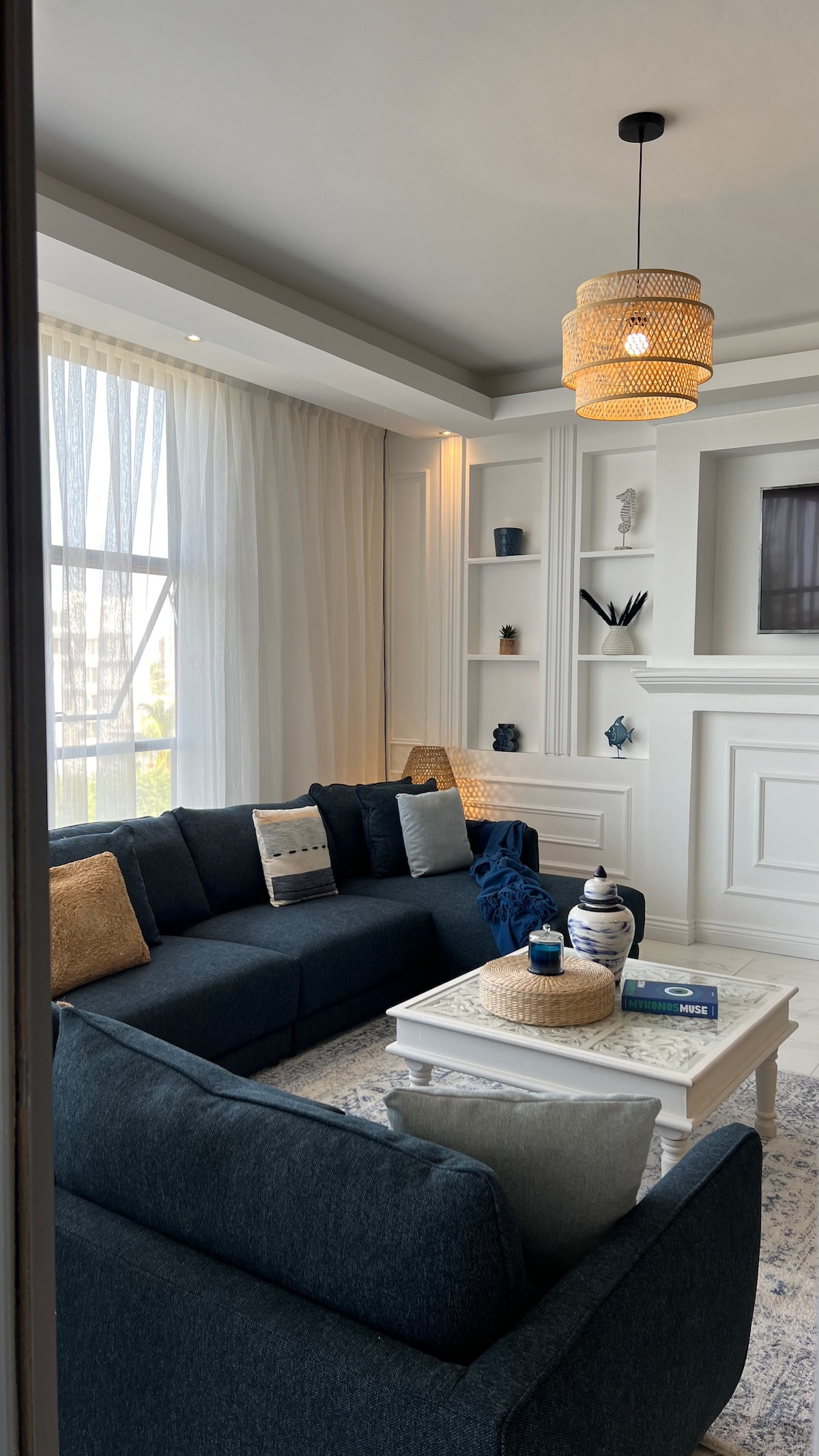 A modern living area is showcased with a dark blue sectional sofa arranged around a light coffee table. Bright curtains frame the large window, allowing natural light to fill the space. Decorative shelves with assorted items add character to the neutral-toned walls.