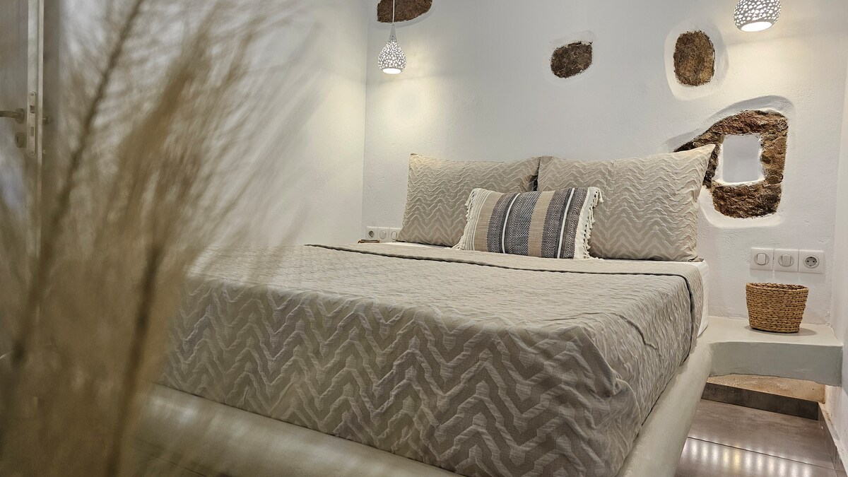 A spacious bedroom features a light-colored patterned bedspread on a large bed. Soft lighting from hanging fixtures complements the natural stone accent wall. Decorative elements include a neutral-toned cushion and a woven basket, creating a serene and inviting sleeping area.