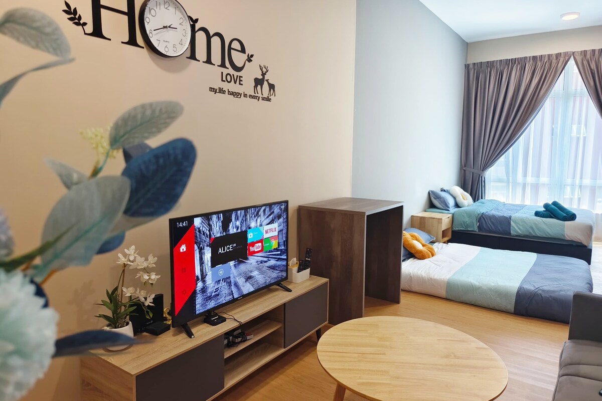 A studio apartment is depicted with a cozy seating area featuring a modern television on a side table. Two spacious beds are arranged in the background, complemented by soft bedding. Natural light streams through large windows, framed by light curtains, enhancing the comfortable ambiance.