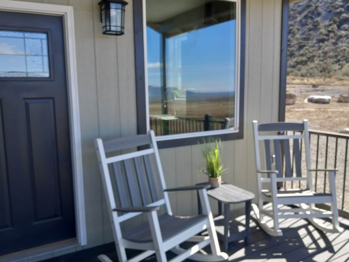 Two rocking chairs are positioned on a welcoming porch, facing a large window that captures the expansive views of the surrounding landscape. A small potted plant sits on the table between the chairs, enhancing the outdoor seating area.