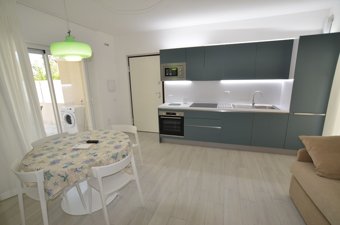 A modern kitchen area is presented, featuring sleek cabinetry and a built-in oven. A round dining table is set with simple decor. Natural light filters through the curtain-adorned sliding doors, leading to an outdoor space. A comfortable sofa complements the layout.