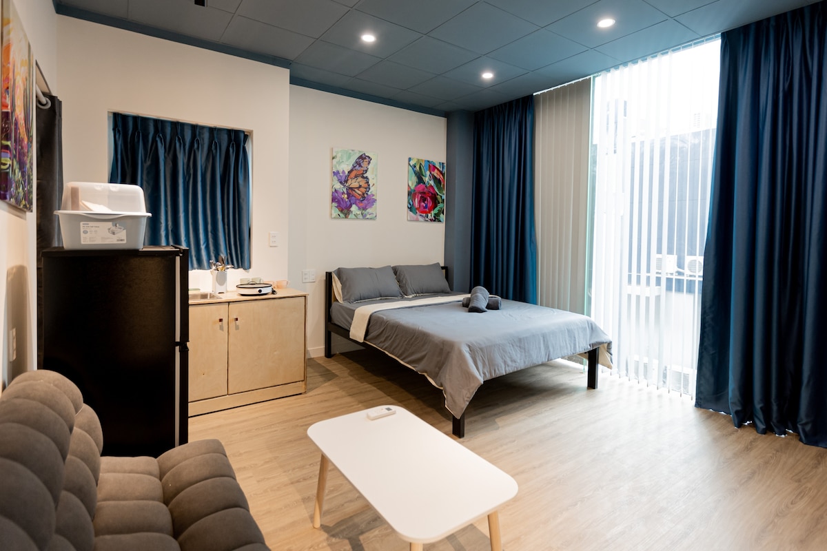A modern studio apartment is presented, featuring a spacious bed with soft linens and two colorful wall art pieces. A well-furnished kitchenette includes a refrigerator, and a comfortable sofa is positioned beside a compact coffee table. Natural light filters through large glass doors adorned with sheer curtains.
