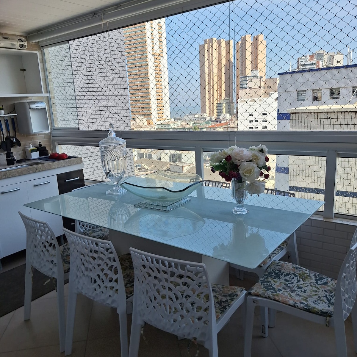 A dining area is presented with a glass table surrounded by six light-colored chairs. A vase filled with flowers and a decorative bowl are placed on the table. Large windows showcase a city view, allowing natural light to enhance the space.