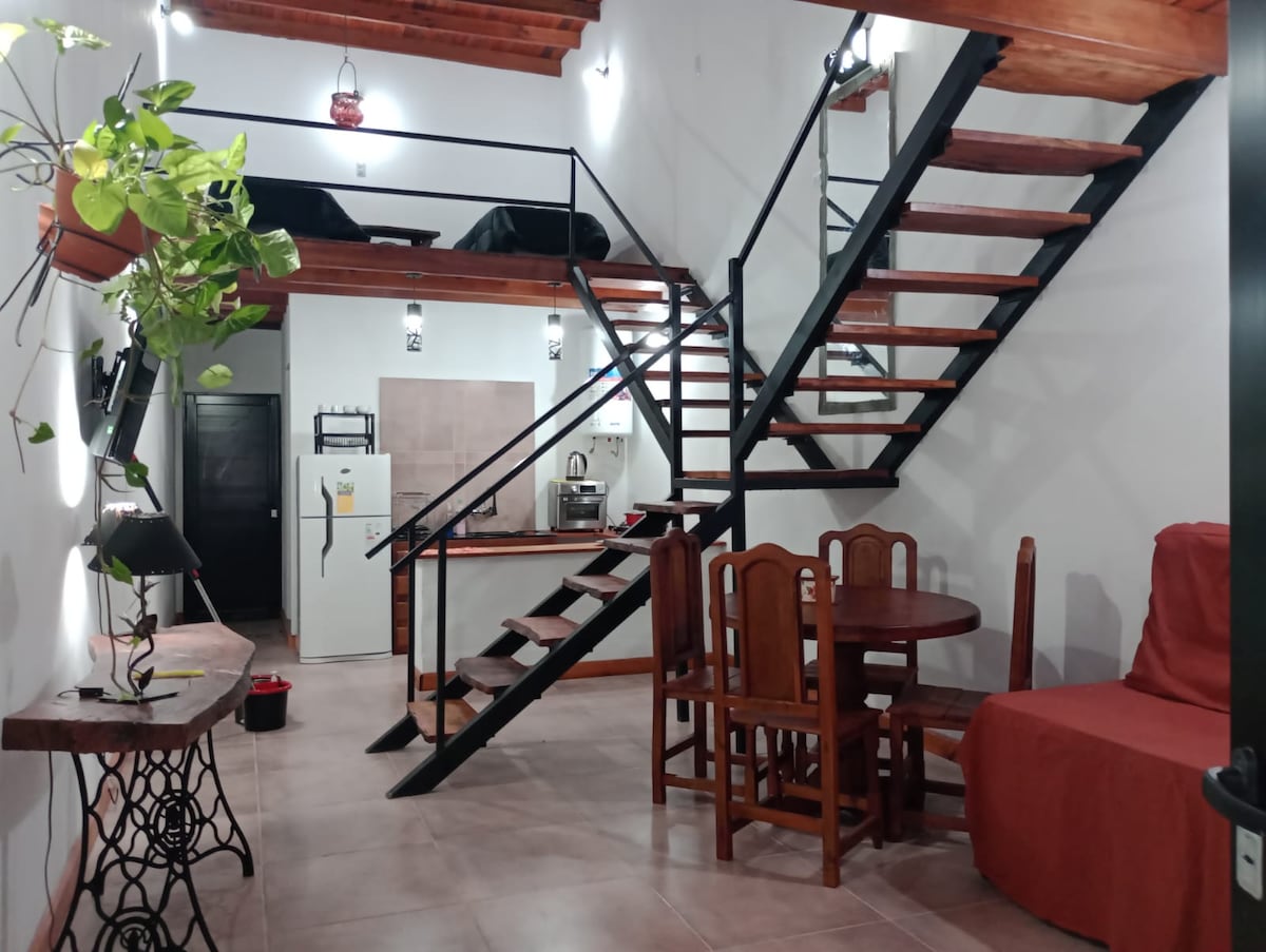 A modern loft interior is presented with an open floor plan, showcasing a staircase with a black metal railing. A wooden dining table with four chairs sits beside a cozy seating area draped in a red cover. A plant adds a touch of greenery to the space.