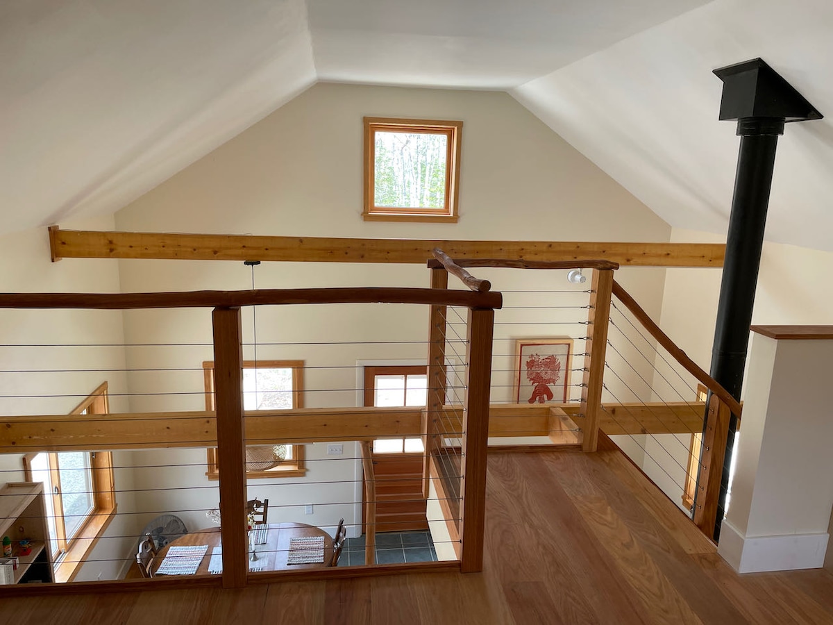 An airy loft area features wooden beams and a central chimney. Natural light streams through a window, illuminating the space. A glimpse of a dining area with a wooden table and chairs can be seen below, enhancing the inviting atmosphere.