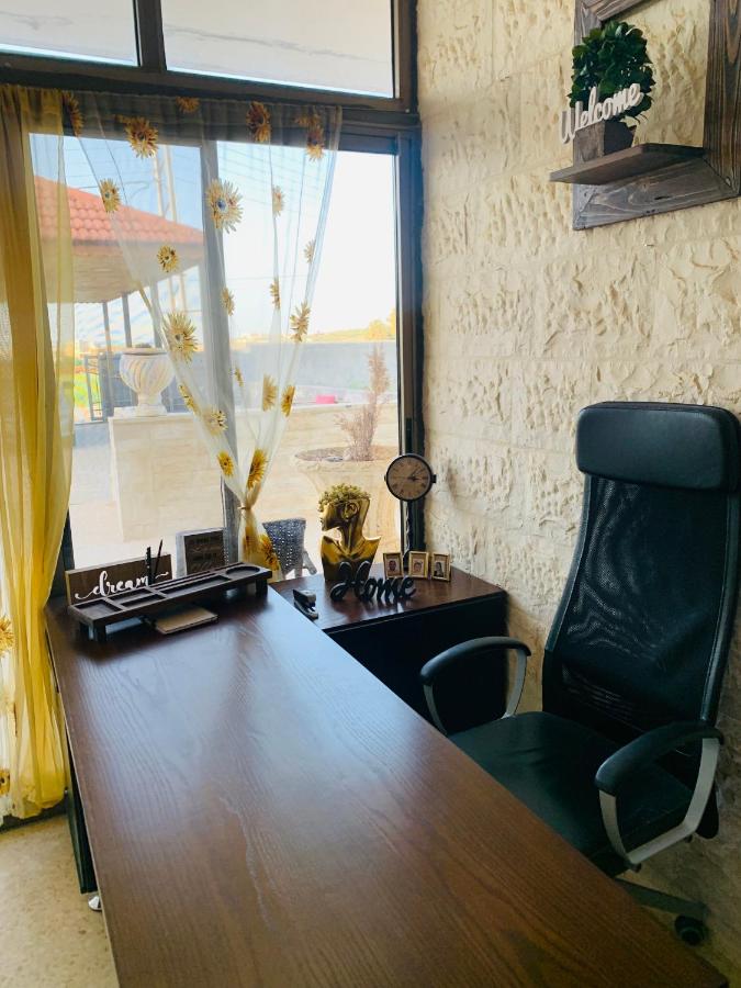 A dedicated workspace features a modern desk topped with decorative items and a wall-mounted shelf displaying a welcoming sign. Sunlight filters through sheer curtains framing a window, creating a bright atmosphere. A comfortable black office chair is positioned ergonomically at the desk.