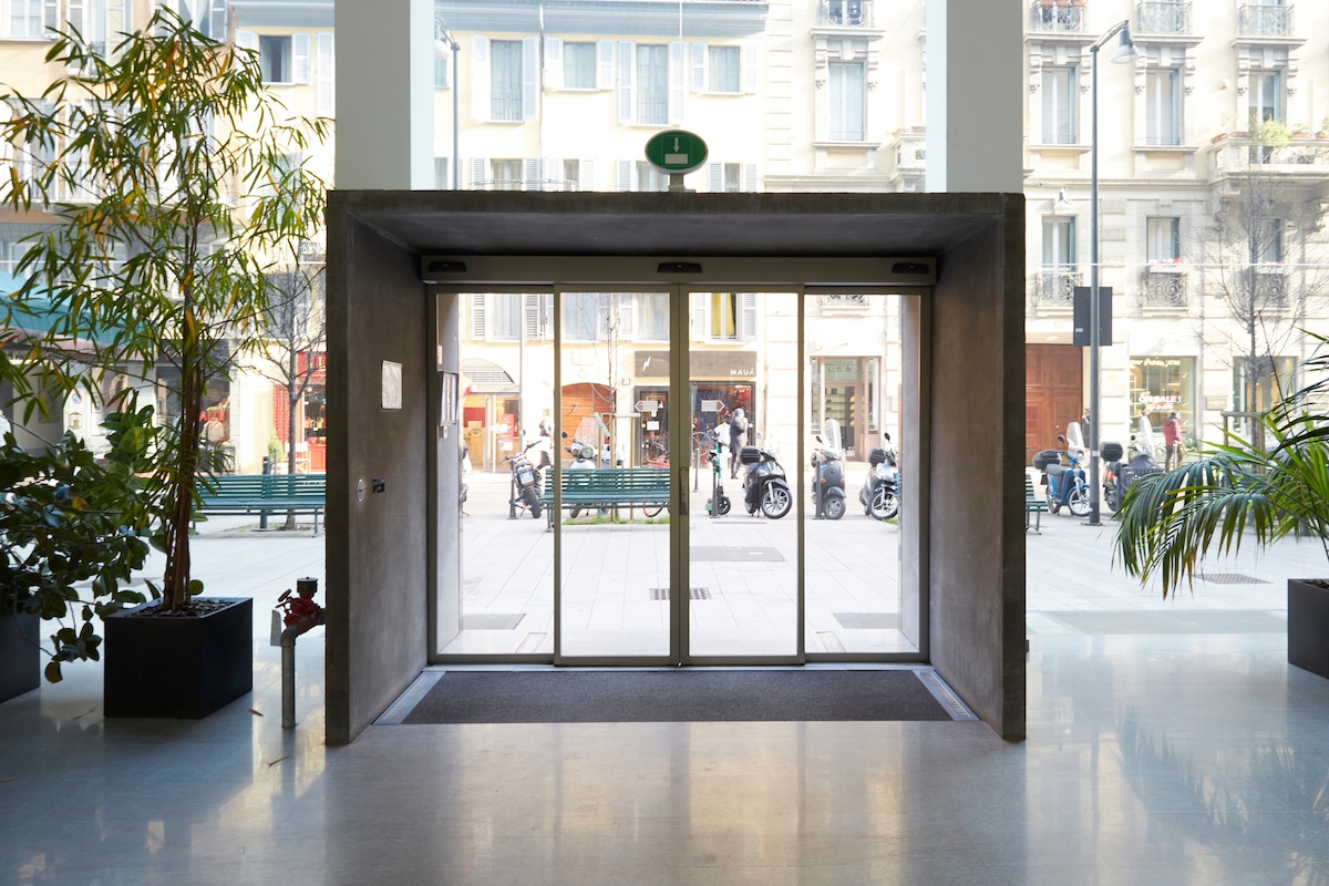 An entrance is framed by large glass doors, allowing natural light to illuminate the space. Potted plants line the interior, and a view of a lively street with benches and parked motorbikes is visible beyond the entrance.