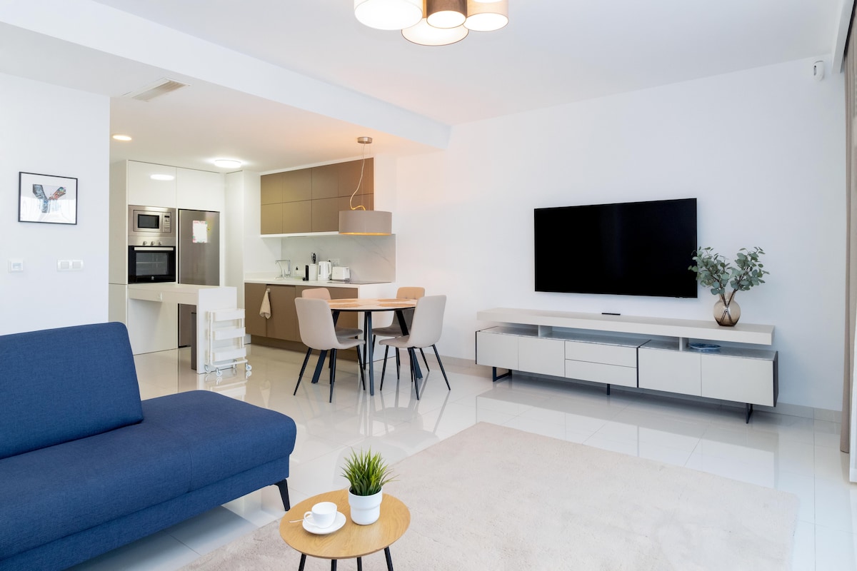 A modern living area features a navy sofa and a light area rug. A flat-screen TV is mounted on the wall opposite a minimalist entertainment unit. An open kitchen with light cabinetry is visible in the background, complemented by a dining table and chairs.