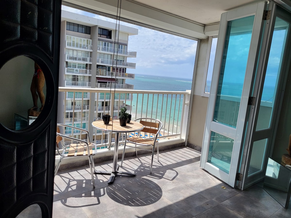 A balcony offers a view of the sea with light reflecting off the water. There is a small round table with two chairs placed near the railing. Glass doors lead to the interior, enhancing the open feel of the space.