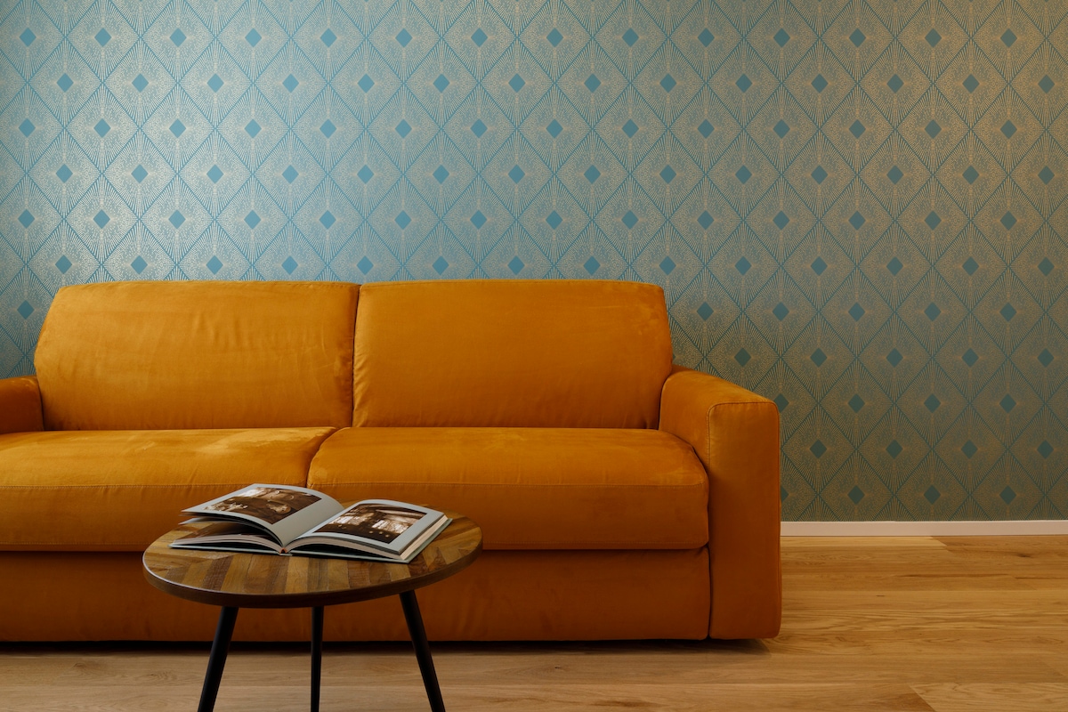 An orange sofa is positioned against a patterned blue wall, creating a bold visual contrast. A wooden side table displays two open magazines, contributing to the modern aesthetic of the living area.