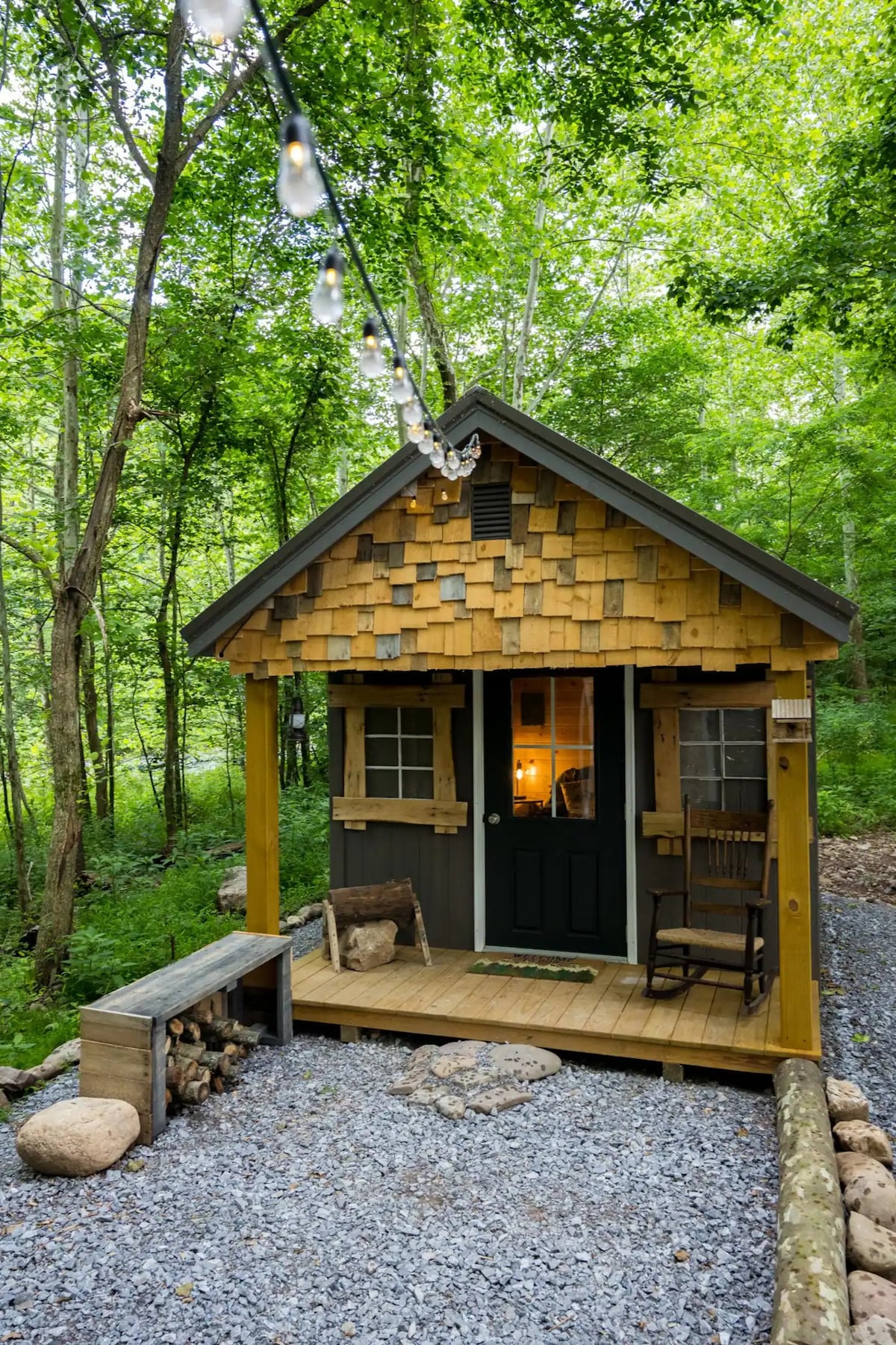 A small cabin is surrounded by trees, featuring a wooden porch with a rocking chair. The exterior showcases a combination of wooden shingles and a green door. Soft lighting from string lights adds a warm ambiance to the outdoor setting.