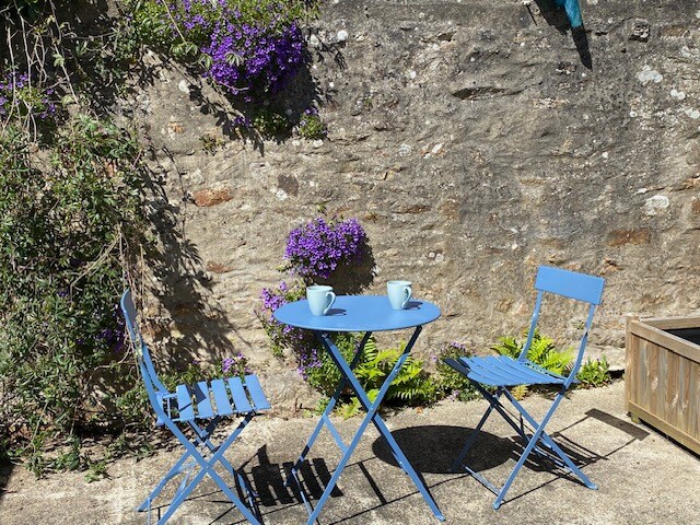A small outdoor seating area features a round blue table accompanied by two matching folding chairs. The backdrop consists of a textured stone wall adorned with vibrant purple flowers, creating a serene ambiance for relaxation.