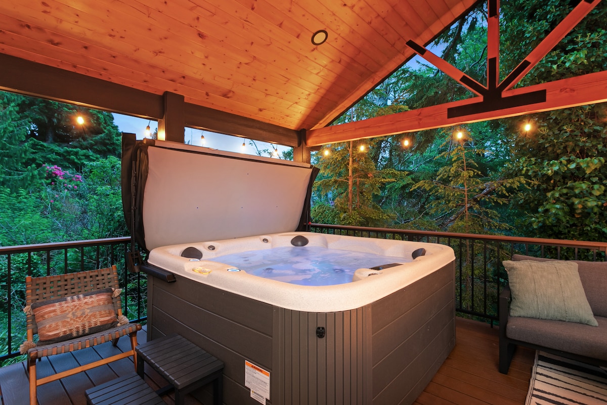 A covered deck features a spacious hot tub, surrounded by lush greenery. Soft lighting is provided by string lights overhead, creating a welcoming ambiance. Outdoor seating includes a chair and small table, enhancing a relaxing outdoor space.