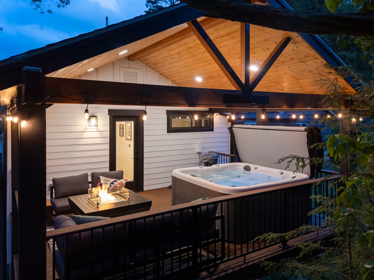 A covered back deck is shown, featuring a hot tub and an outdoor sectional sofa arranged around a propane fire table. Soft lighting illuminates the space, emphasizing wooden beams and a cozy atmosphere. Access to the home is visible through a nearby door.