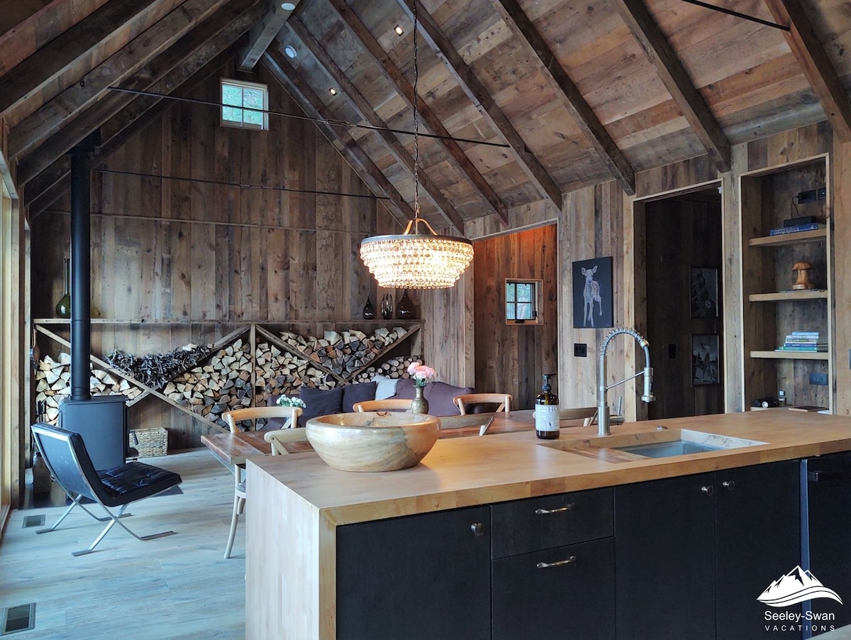 An open kitchen and dining space features wooden walls and a vaulted ceiling, creating a warm ambiance. A modern chandelier hangs above a large island, showcasing a bowl and wine bottle. Cozy seating is arranged around a stack of firewood, enhancing the inviting atmosphere.