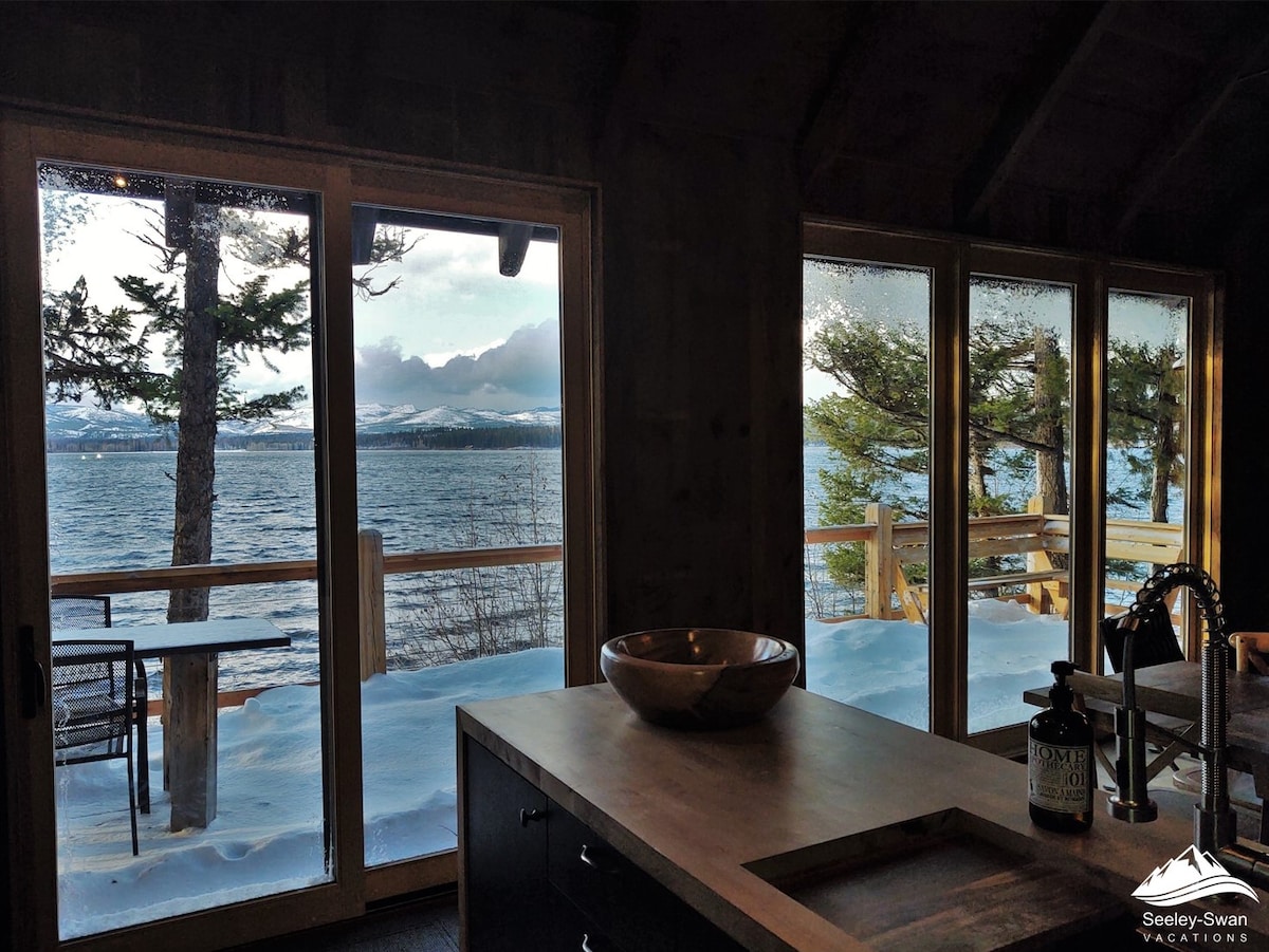 Large glass doors frame a stunning view of the snow-covered deck and Placid Lake, inviting natural light into the open interior. The scene captures the calm water and distant mountains, while tall pine trees are visible through the windows.