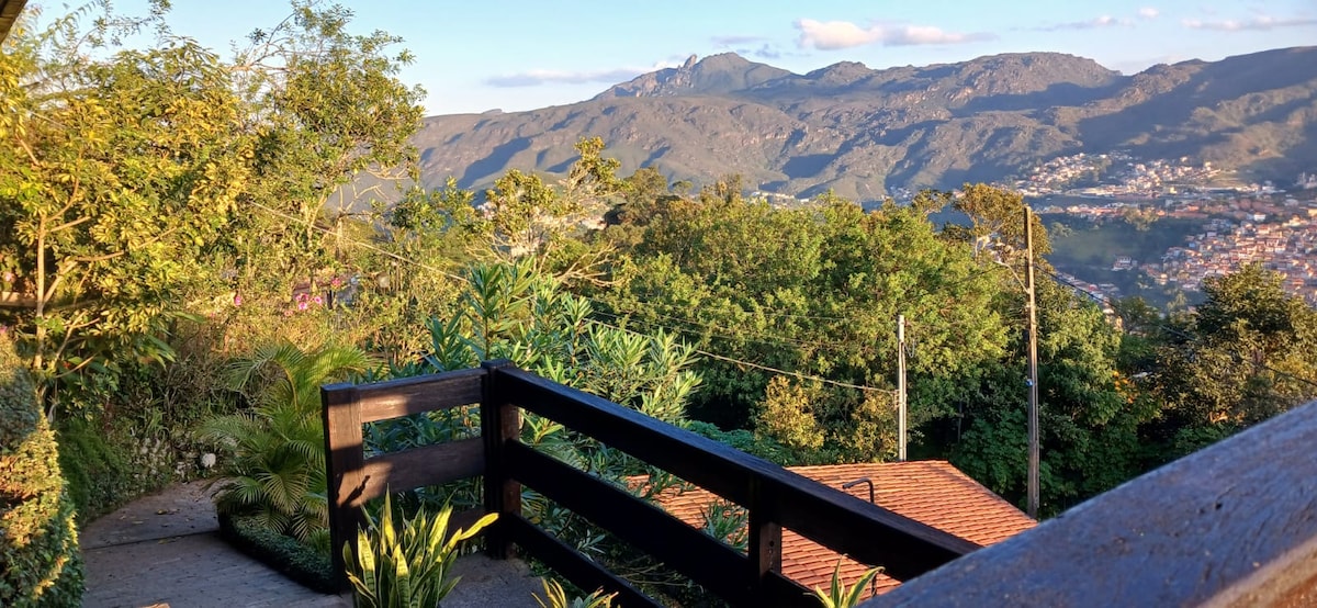 A balcony railing frames a serene view of lush greenery and distant mountains. The landscape features rolling hills under a clear sky, accented by scattered clouds. Nearby, tropical plants add natural beauty, enhancing the outdoor space's connection to the surrounding environment.
