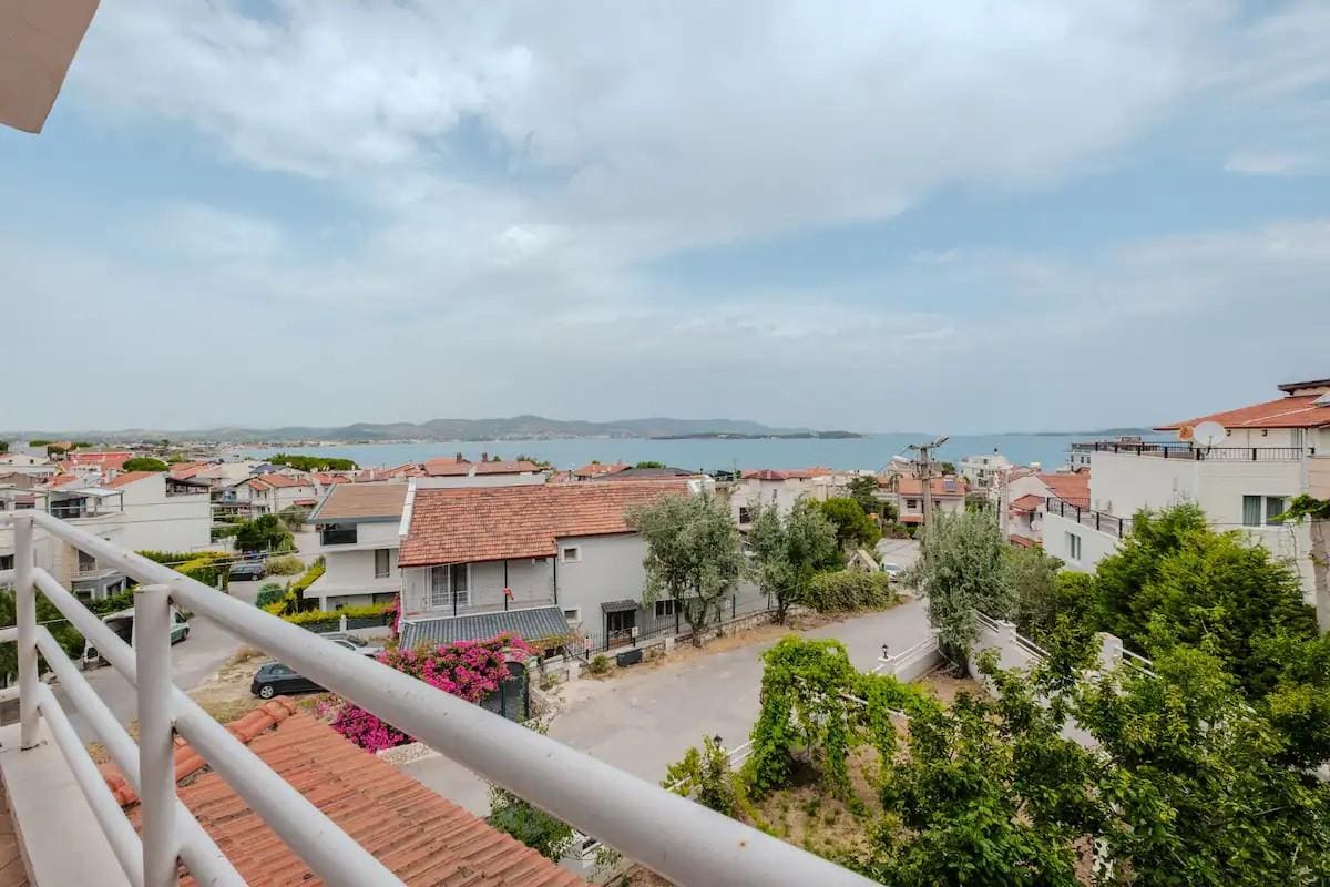 A balcony offers a sweeping view of the surrounding residential area and the sea in the distance. The landscape features various rooftops, greenery, and a cloudy sky, creating a serene backdrop for relaxing outdoors.