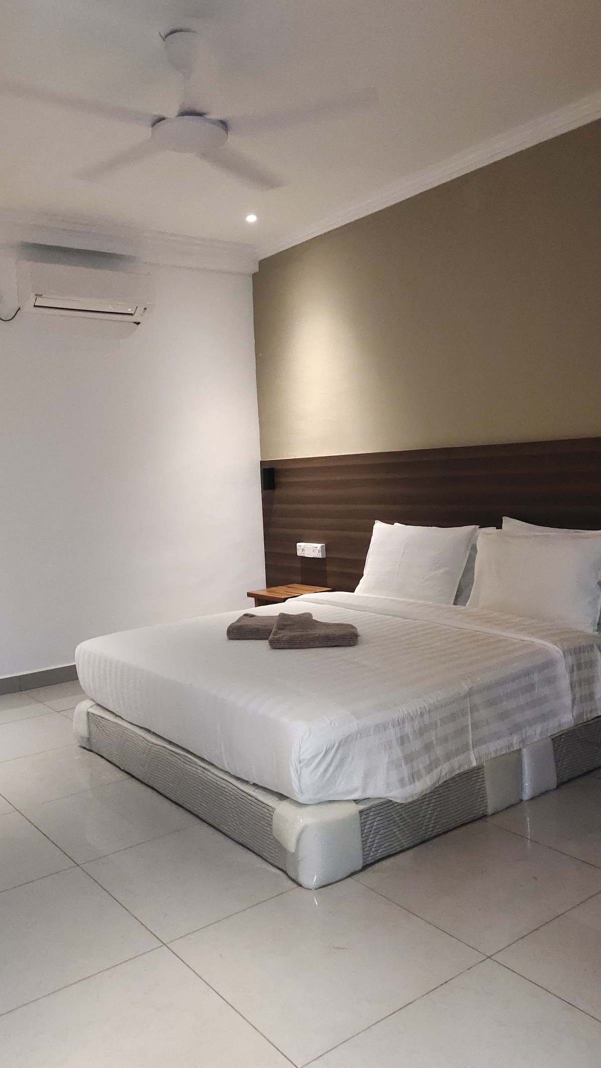 A spacious room features a large king-sized bed with crisp linens and two folded towels resting on the bed. The walls are painted in soft tones, complemented by a wooden headboard. Natural light is welcomed through the ceiling-mounted fan and a nearby air conditioning unit.